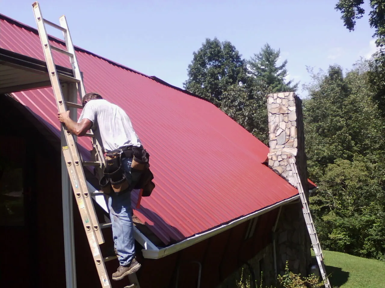 Roofer on a ladder beside a red metal roof and stone chimney — Soffit & Fascia Repair by Roofing Brothers in Tremont City